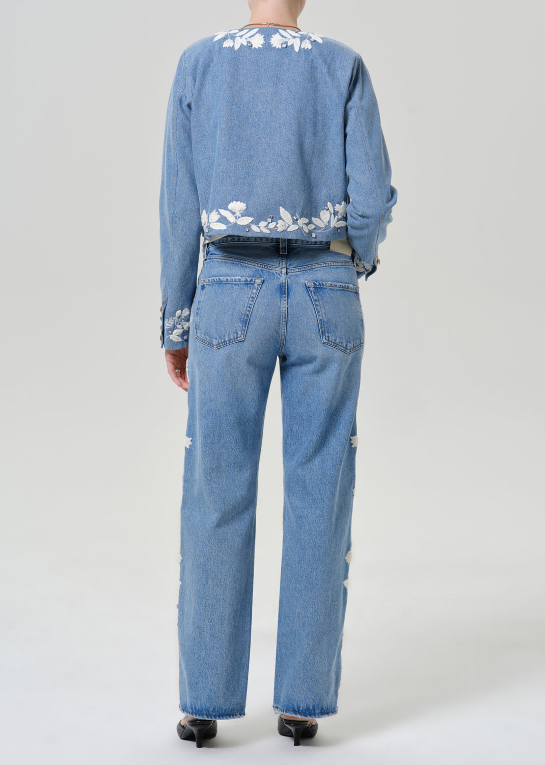 Woman wearing light blue floral jeans with a matching light blue floral denim jacket viewed from the back on a grey background.