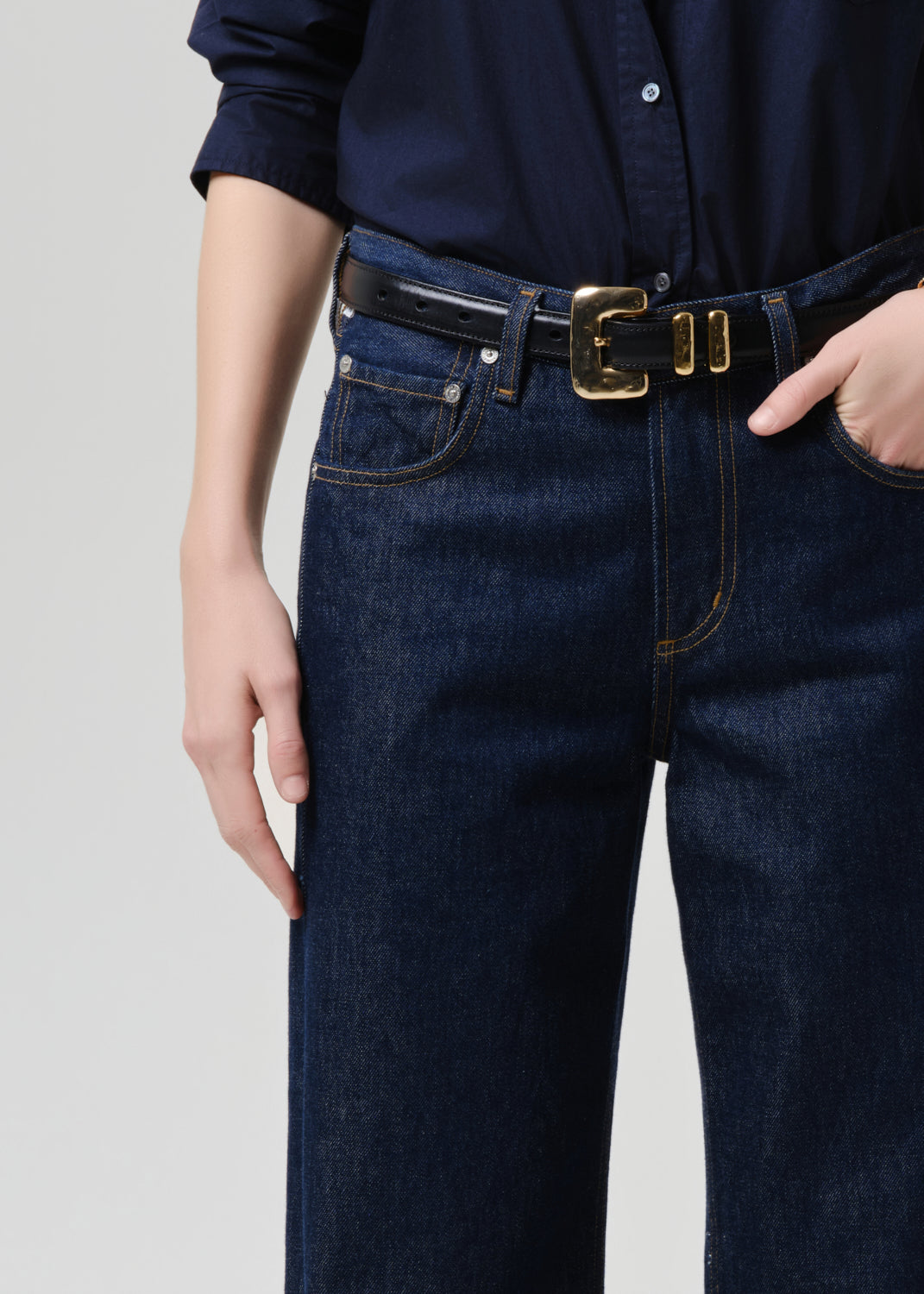 Close-up of a person wearing dark blue denim jeans with a black leather belt featuring a prominent gold-tone double-buckle, paired with a navy shirt against a grey background.