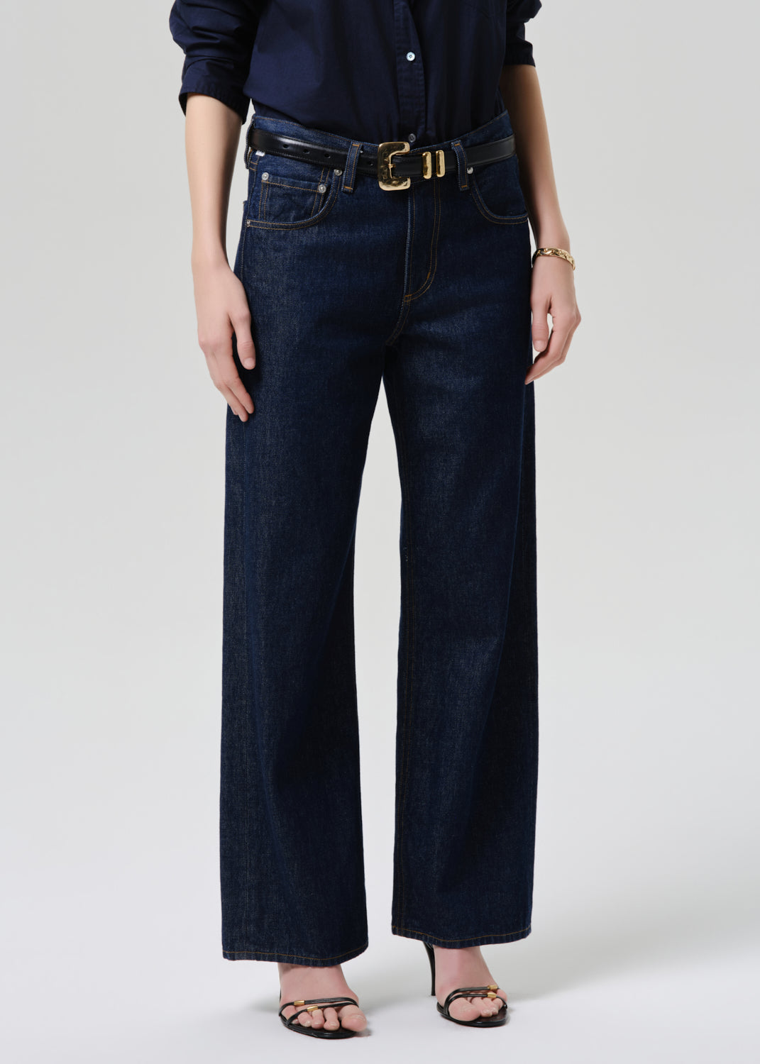 A model wearing dark blue wide-leg denim jeans with a black belt featuring a gold buckle, styled with a navy shirt and black sandals against a grey background.