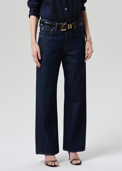 A model wearing dark blue wide-leg denim jeans with a black belt featuring a gold buckle, styled with a navy shirt and black sandals against a grey background.