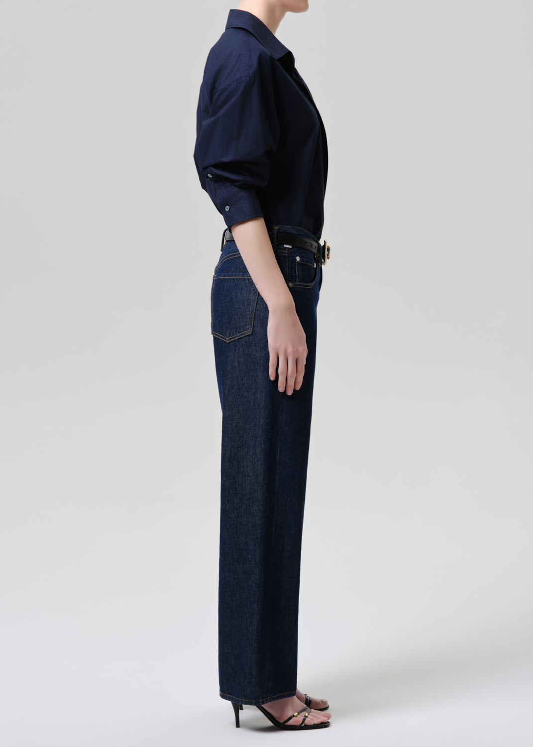 A side-profile shot of a model wearing dark blue wide-leg jeans, a navy long-sleeve button-up shirt, and dark brown heeled sandals against a grey background.