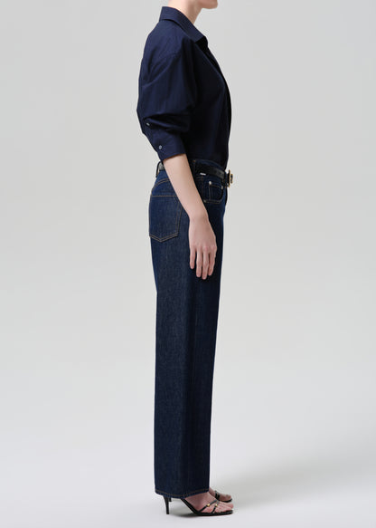 A side-profile shot of a model wearing dark blue wide-leg jeans, a navy long-sleeve button-up shirt, and dark brown heeled sandals against a grey background.