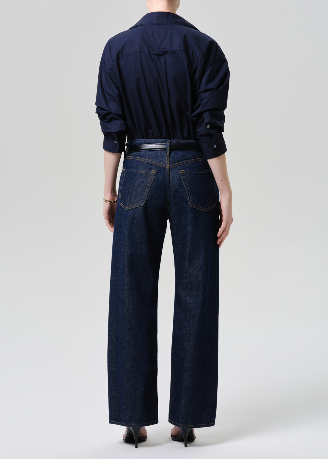 Rear view of a model wearing a dark navy blue long-sleeve button-up shirt tucked into matching dark blue wide-leg jeans, styled with a thin black belt and black heels against a grey background.