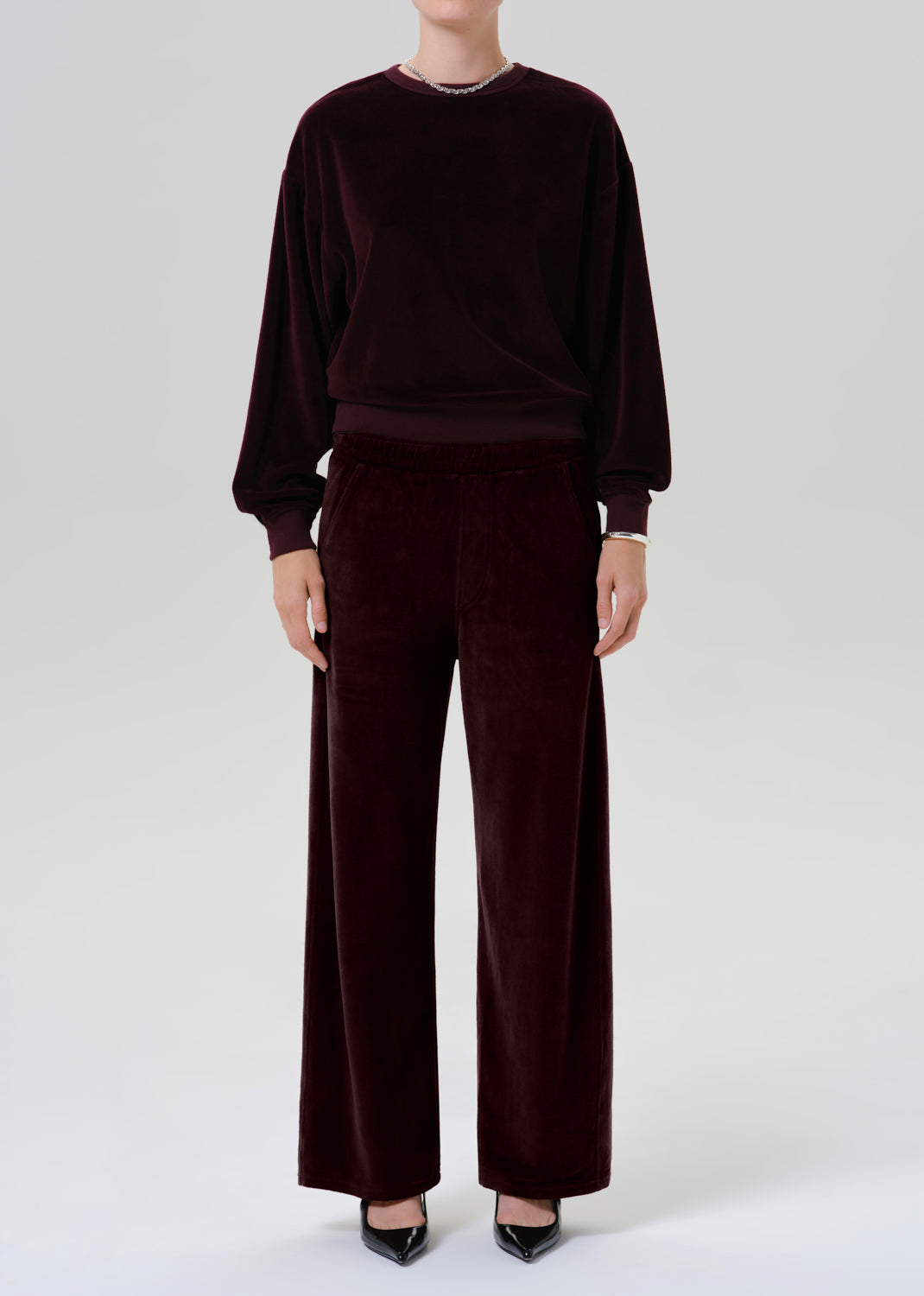 A model wearing a matching dark burgundy velvet lounge set, consisting of a long-sleeve sweatshirt and wide-leg trousers, paired with black pointed-toe heels against a grey background.