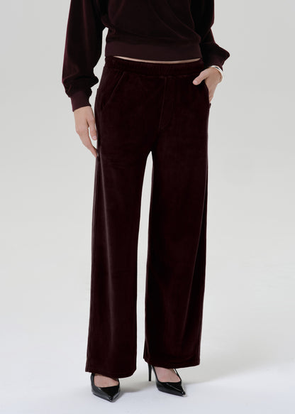A model wearing a matching deep burgundy velvet lounge set, featuring wide-leg pants and a long-sleeve top, paired with black pointed-toe heels against a grey background.
