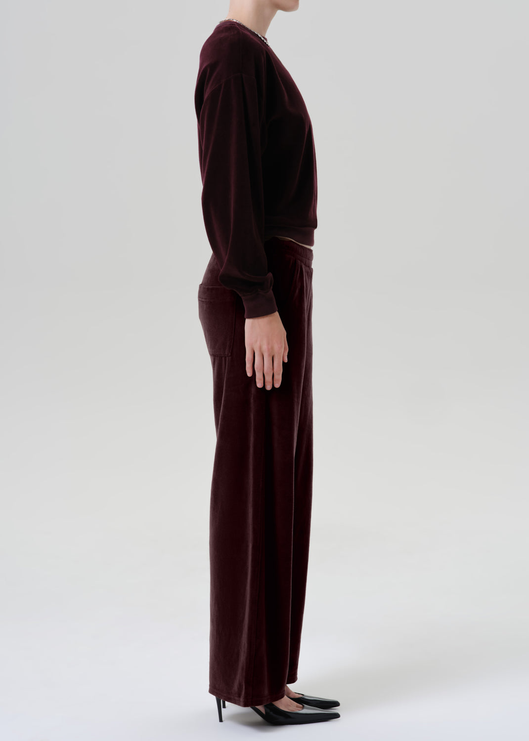 A model shown from the side wearing a matching dark burgundy velvet long-sleeve top and wide-leg trousers set, paired with black pointed-toe flats, against a grey background.