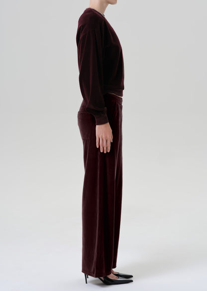 A model shown from the side wearing a matching dark burgundy velvet long-sleeve top and wide-leg trousers set, paired with black pointed-toe flats, against a grey background.