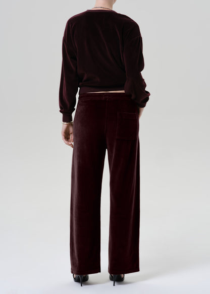 A model shown from the back wearing a matching deep burgundy velvet lounge set, consisting of a long-sleeve cropped top and wide-leg trousers, paired with black high heels against a grey background.