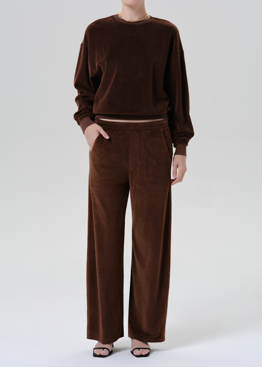 A model wearing a matching dark brown lounge set consisting of a long-sleeve crewneck sweatshirt and wide-leg trousers, styled with black flat sandals against a grey background.