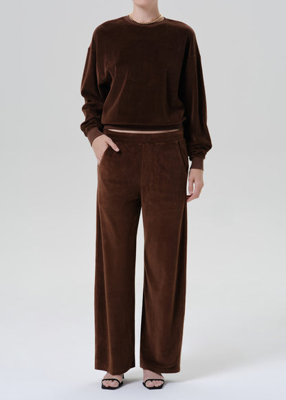 A model wearing a matching dark brown lounge set consisting of a long-sleeve crewneck sweatshirt and wide-leg trousers, styled with black flat sandals against a grey background.