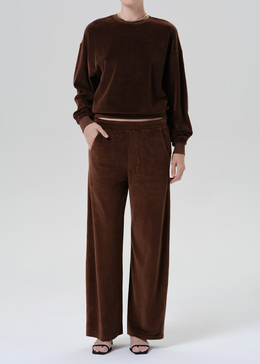 A model wearing a matching dark brown lounge set consisting of a long-sleeve crewneck sweatshirt and wide-leg trousers, styled with black flat sandals against a grey background.