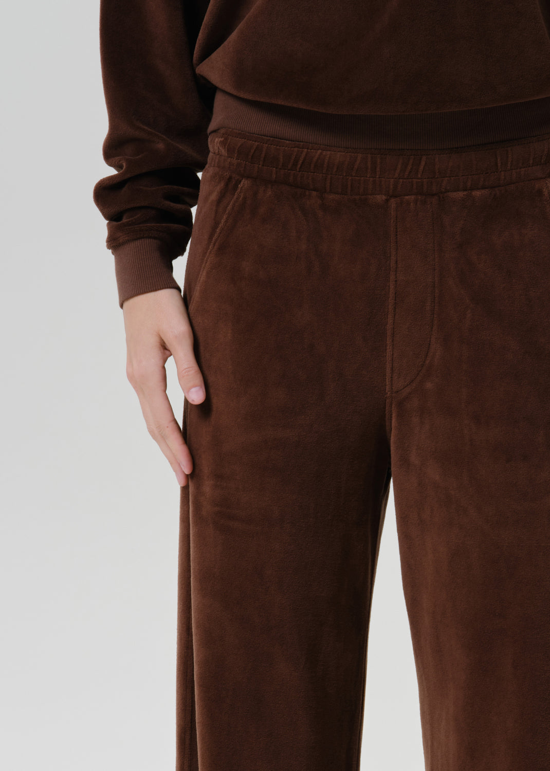 A close-up of a person wearing dark brown velvet lounge pants and a matching long-sleeve top against a grey background.