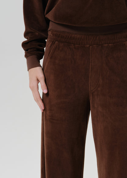 A close-up of a person wearing dark brown velvet lounge pants and a matching long-sleeve top against a grey background.