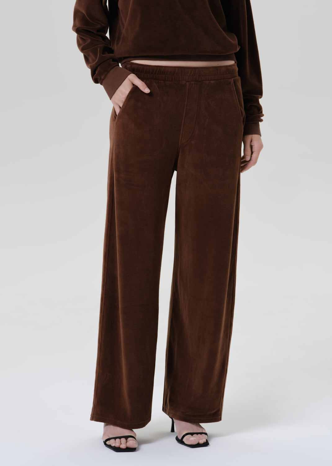 A model wearing a matching brown velvet lounge set, featuring wide-leg trousers and a cropped long-sleeve top, paired with black heeled sandals against a grey background.