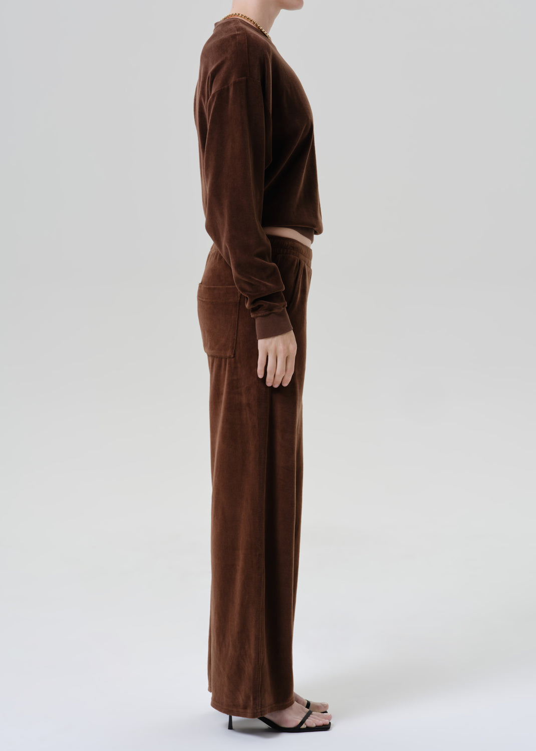 A model shown from the side wearing a matching brown corduroy long-sleeve crop top and wide-leg trousers set, paired with black heeled sandals against a grey background.