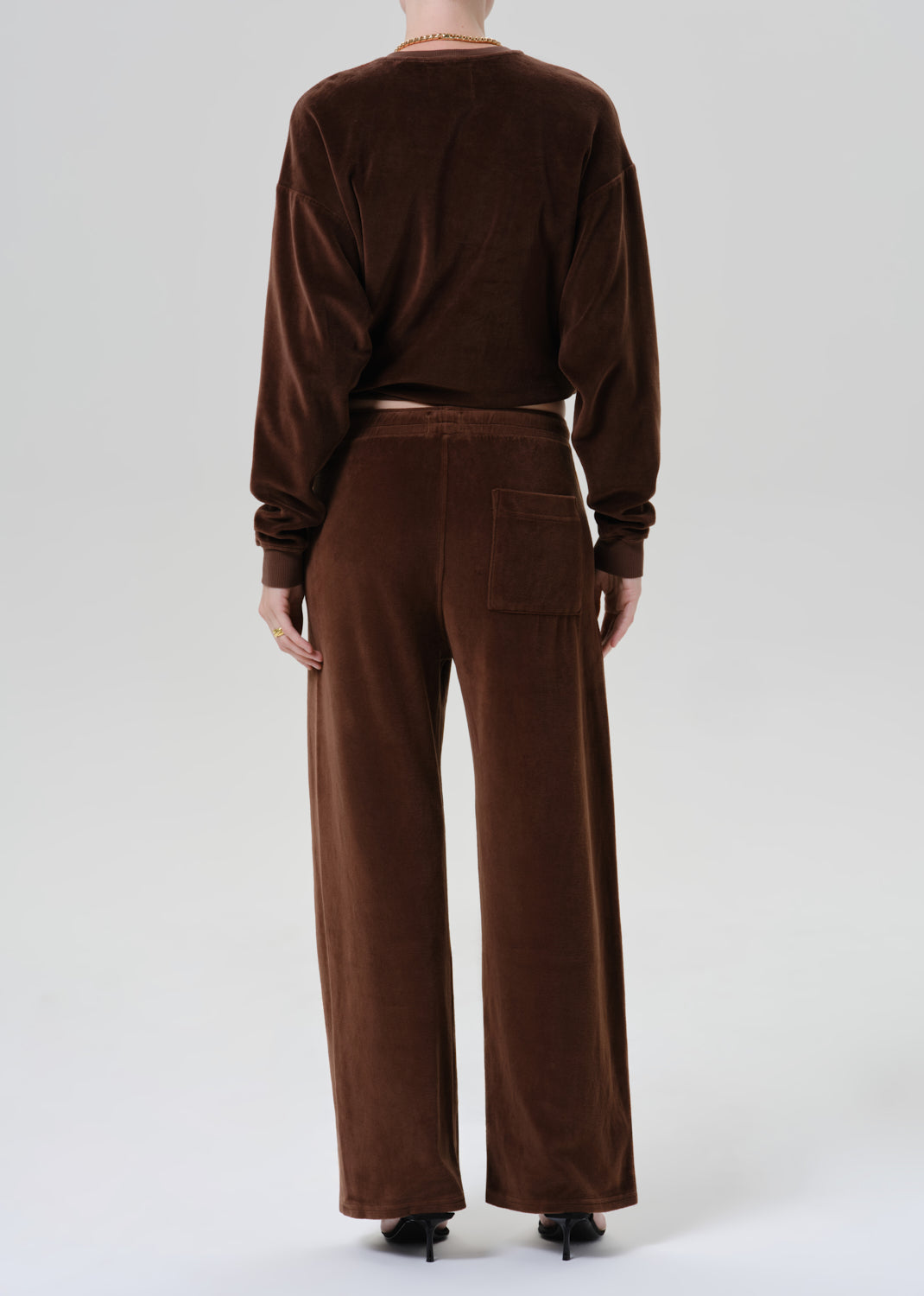 A model viewed from the back wearing a matching brown velvet long-sleeve crop top and wide-leg trousers set, paired with black pointed-toe heels against a grey background.
