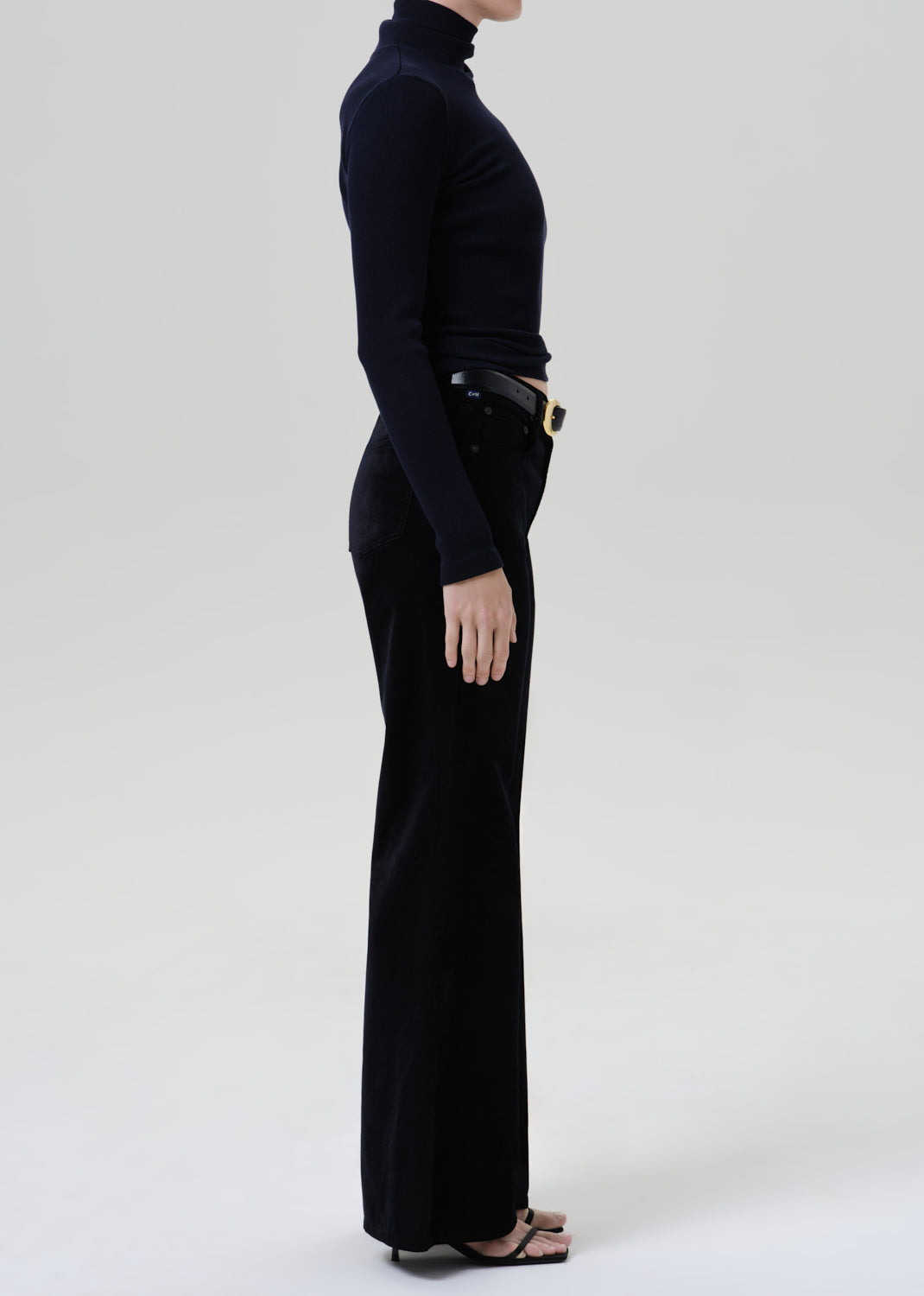 Person wearing a black fitted turtleneck with black wide leg trousers viewed from the side on a grey background
