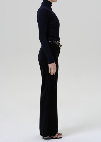 Person wearing a black fitted turtleneck with black wide leg trousers viewed from the side on a grey background