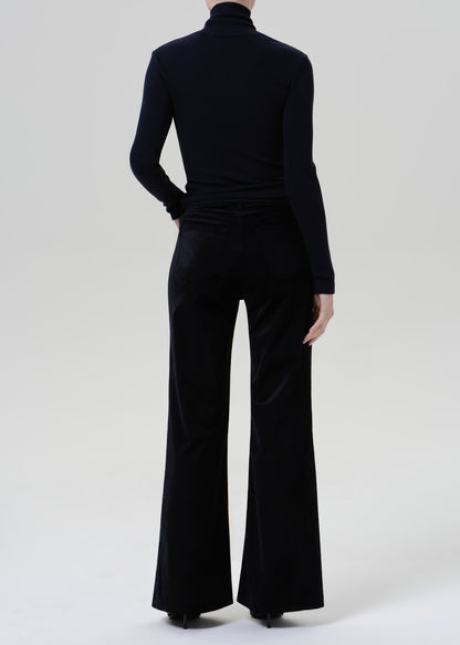 Person wearing a black fitted turtleneck with black wide leg trousers viewed from the back on a grey background