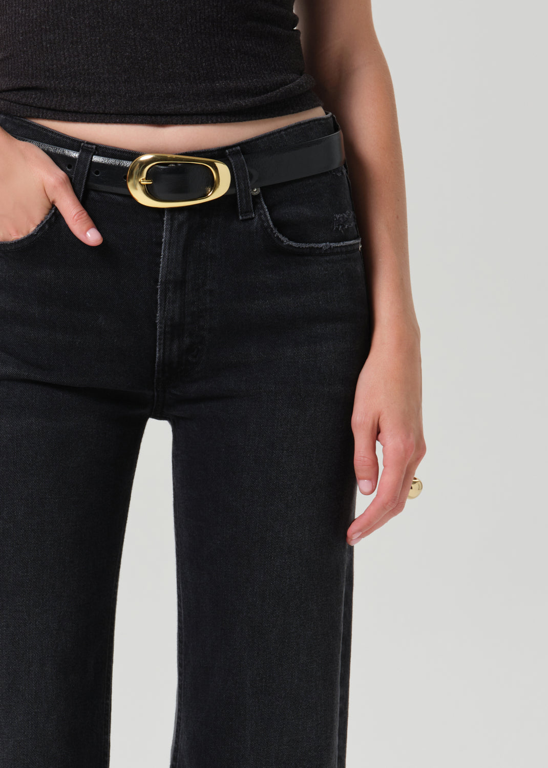 Close-up of a model wearing black jeans with a black leather belt featuring a prominent oval gold-tone buckle, paired with a black ribbed crop top against a grey background.