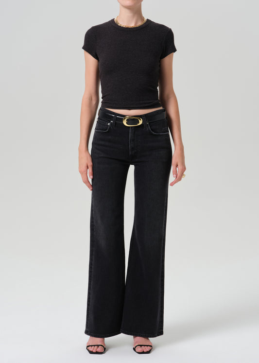 A model wearing a black short-sleeve crop top and black wide-leg jeans with a gold-tone belt buckle, standing against a grey background.
