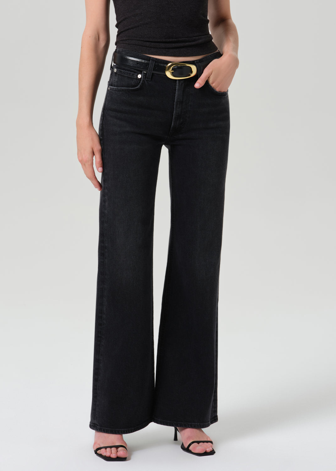 A model wearing black wide-leg jeans with a black leather belt featuring a gold oval buckle, paired with a black crop top and black heeled sandals, against a grey background.