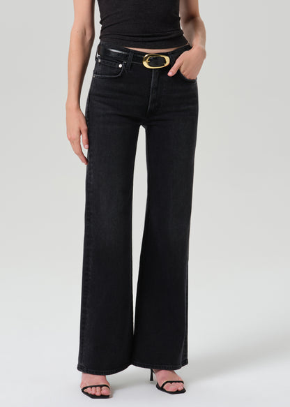 A model wearing black wide-leg jeans with a black leather belt featuring a gold oval buckle, paired with a black crop top and black heeled sandals, against a grey background.
