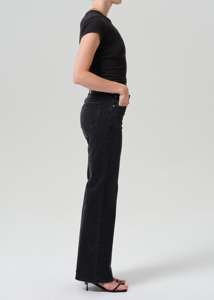 A side-profile studio shot of a model wearing black wide-leg jeans, a black short-sleeve top, and black heeled sandals against a grey background.