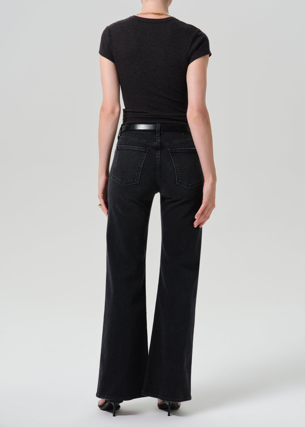 A model shown from the back wearing a black short-sleeve top and black wide-leg jeans with a thin black belt, photographed against a grey background.