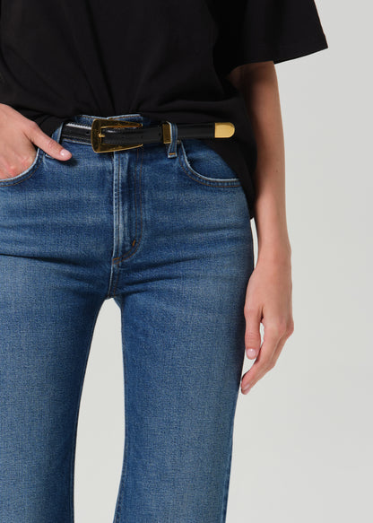 A close-up of a person wearing blue jeans with a black belt featuring a gold-tone buckle and tip, paired with a black top against a grey background.