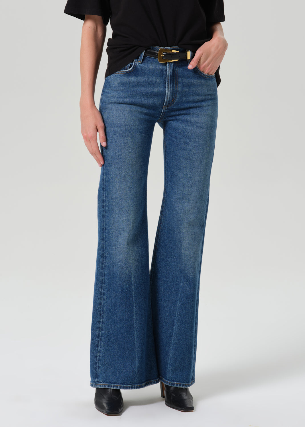A model wearing a pair of medium-wash blue flare jeans with a black top and black boots, shown from the waist down against a grey background.