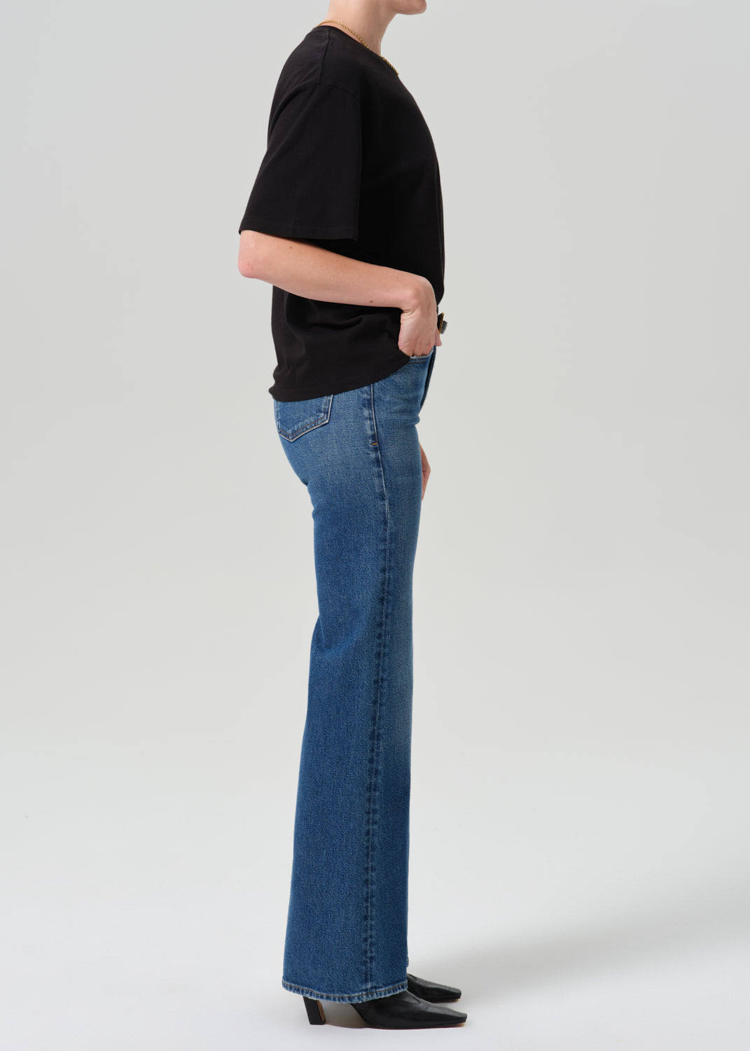 A side-profile studio shot of a model wearing blue flare jeans, a black short-sleeve t-shirt, and black pointed-toe heels against a grey background.