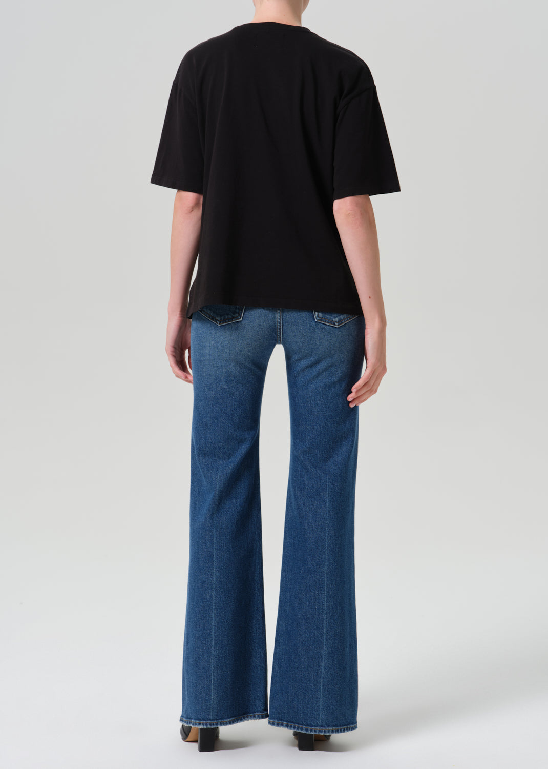 A model shown from the back wearing a black short-sleeve t-shirt and blue wide-leg flare jeans against a grey background.