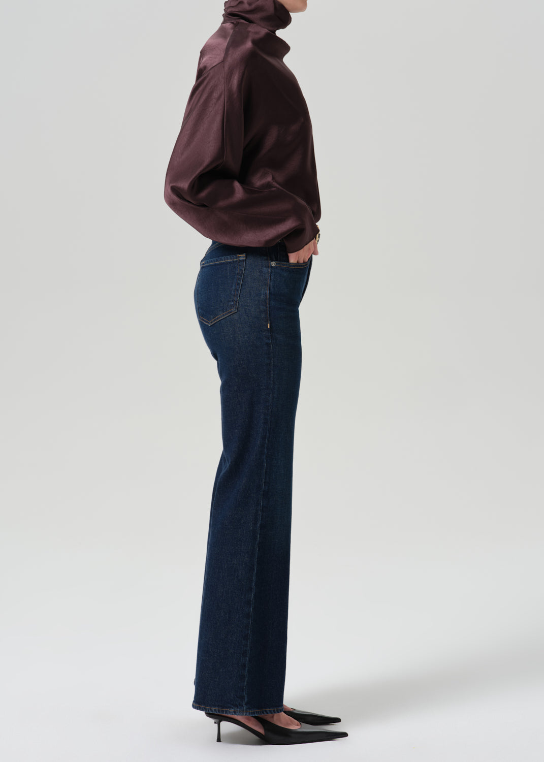 Person wearing a dark brown long sleeve draped blouse with dark indigo cropped wide leg jeans viewed from the side on a grey background