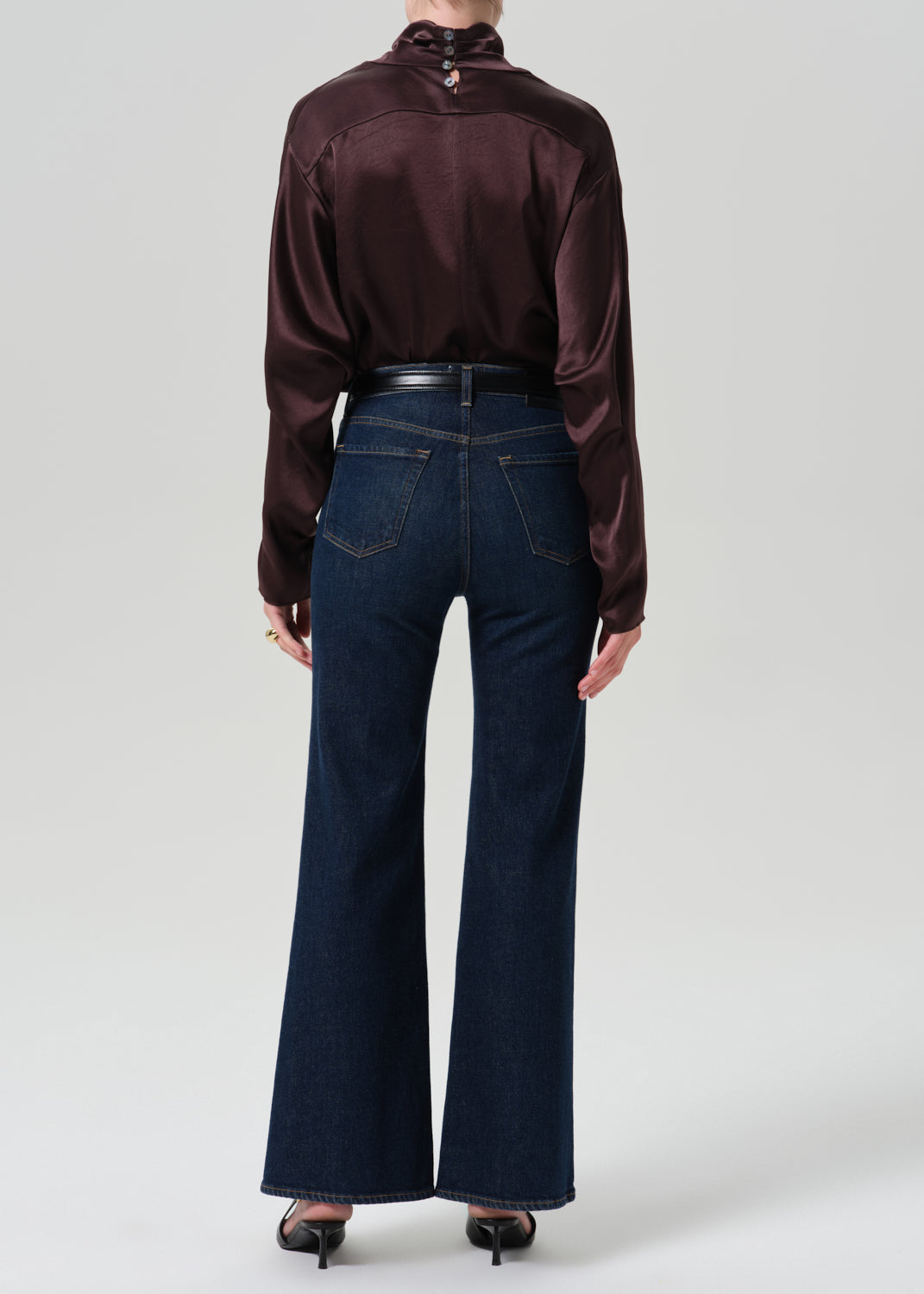 Person wearing a dark brown long sleeve draped blouse with dark indigo cropped wide leg jeans viewed from the back on a grey background