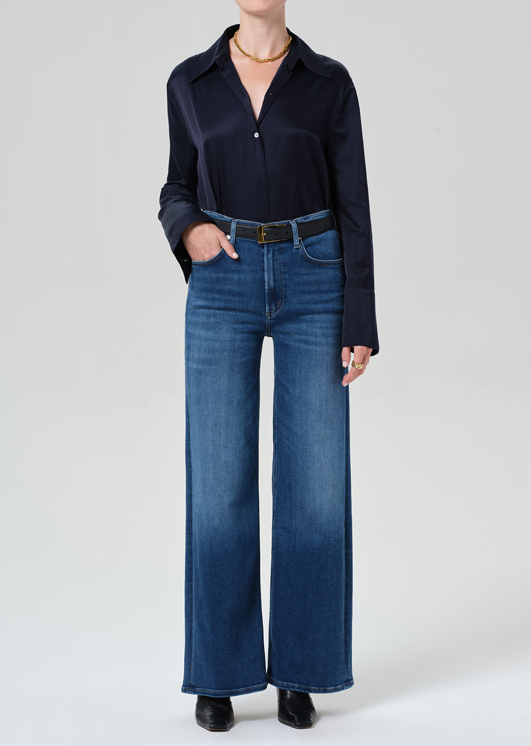 A model wearing a dark navy long-sleeve button-up shirt and high-waisted, wide-leg blue jeans with a black belt, standing against a grey background.