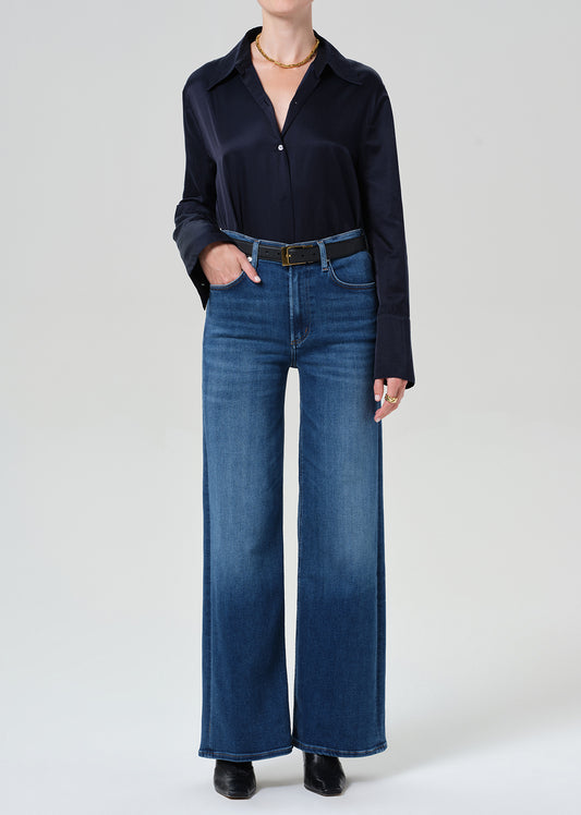A model wearing a dark navy long-sleeve button-up shirt and high-waisted, wide-leg blue jeans with a black belt, standing against a grey background.