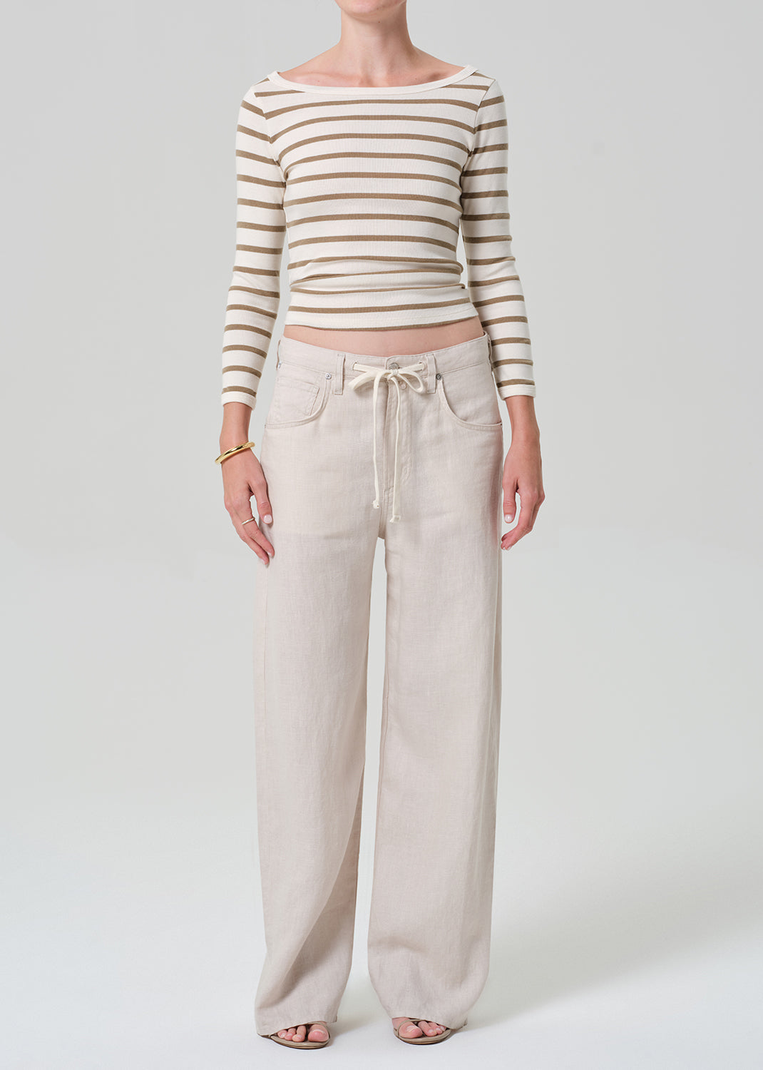 A model wearing a beige and brown horizontally striped long-sleeve crop top and light beige wide-leg linen pants with a drawstring waist, standing against a grey background.