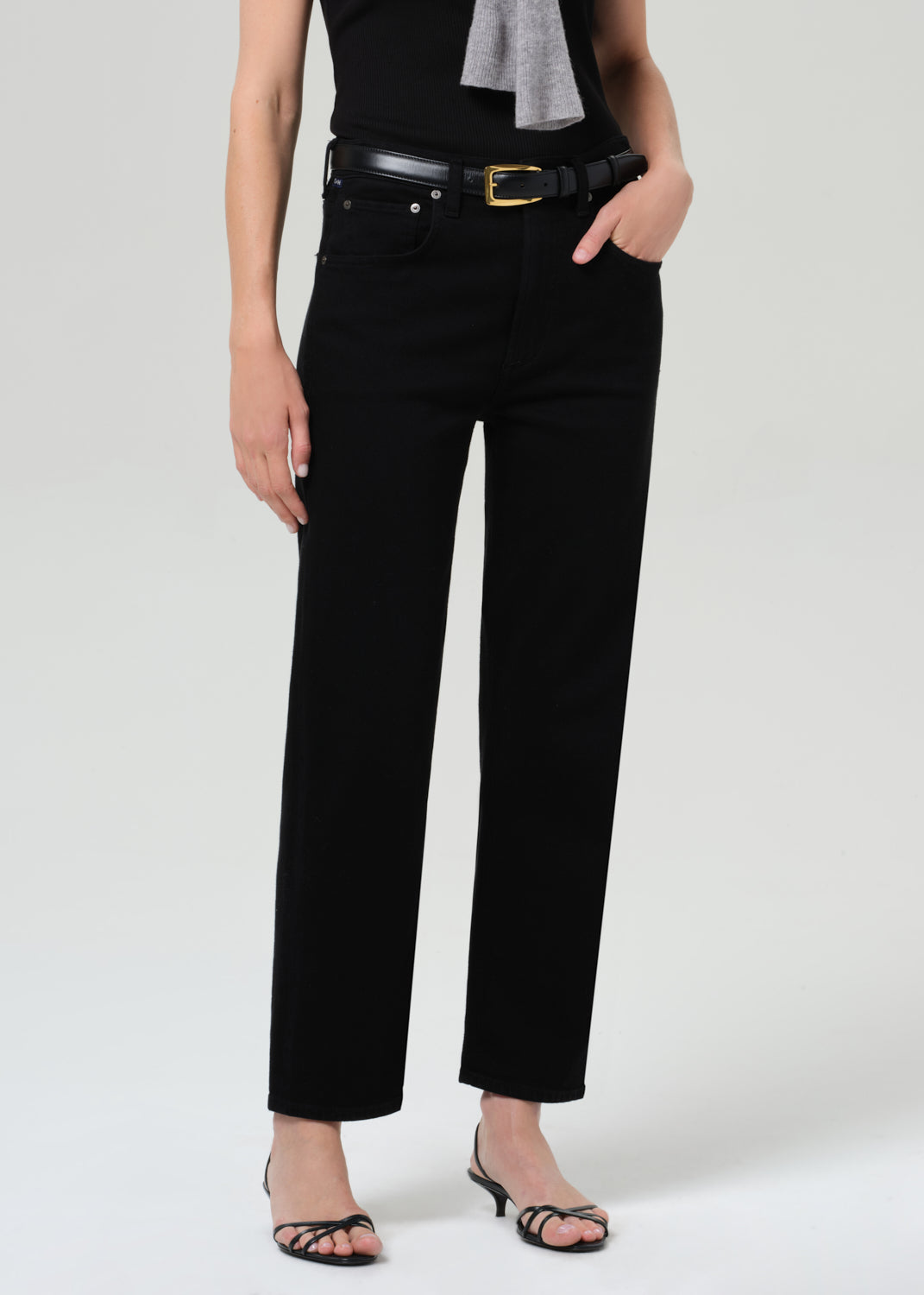 Person wearing a black sleeveless top and pants with a gray sweater, standing against a grey background