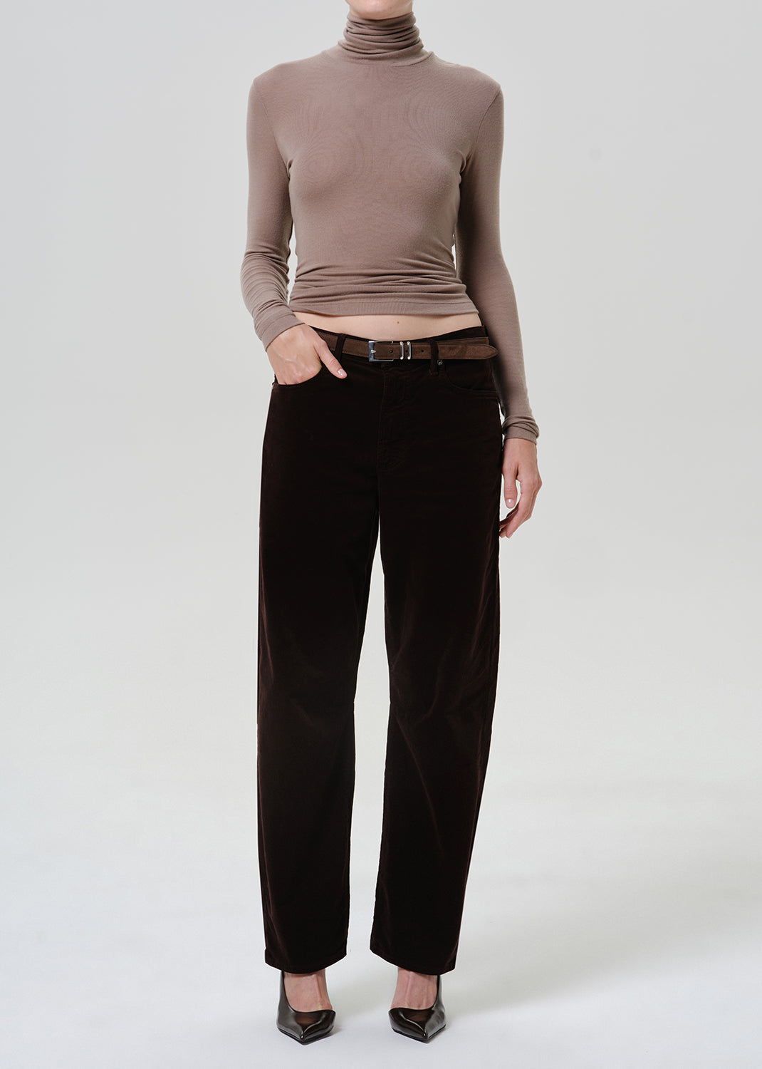 A model wearing a taupe-brown ribbed turtleneck top and dark brown wide-leg trousers, styled with a brown belt and black pointed-toe heels against a light gray studio background.