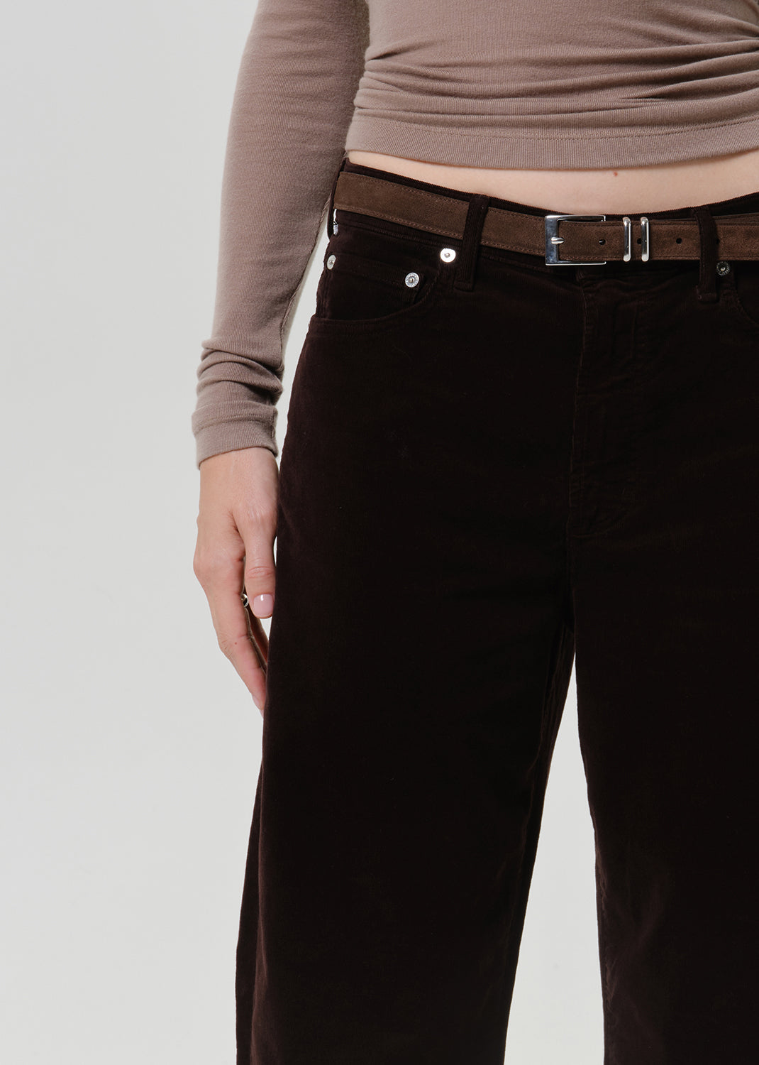 A close-up of a person wearing dark brown wide-leg corduroy pants with a brown belt and silver buckle, paired with a taupe long-sleeve top, against a light gray studio background.
