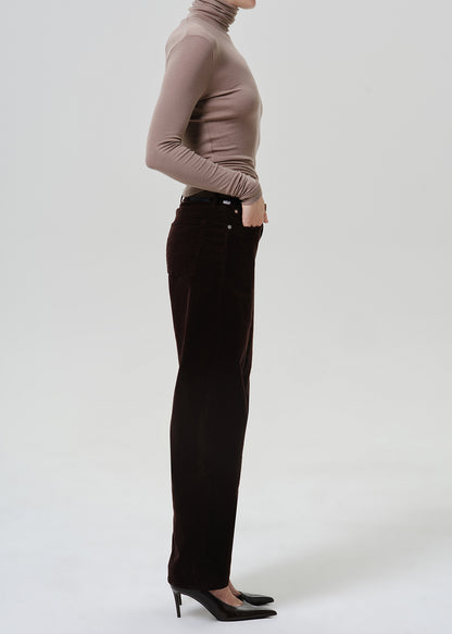 A side-profile studio shot of a model wearing dark brown corduroy trousers, a taupe-brown ribbed turtleneck, and black pointed-toe heels against a light gray background.