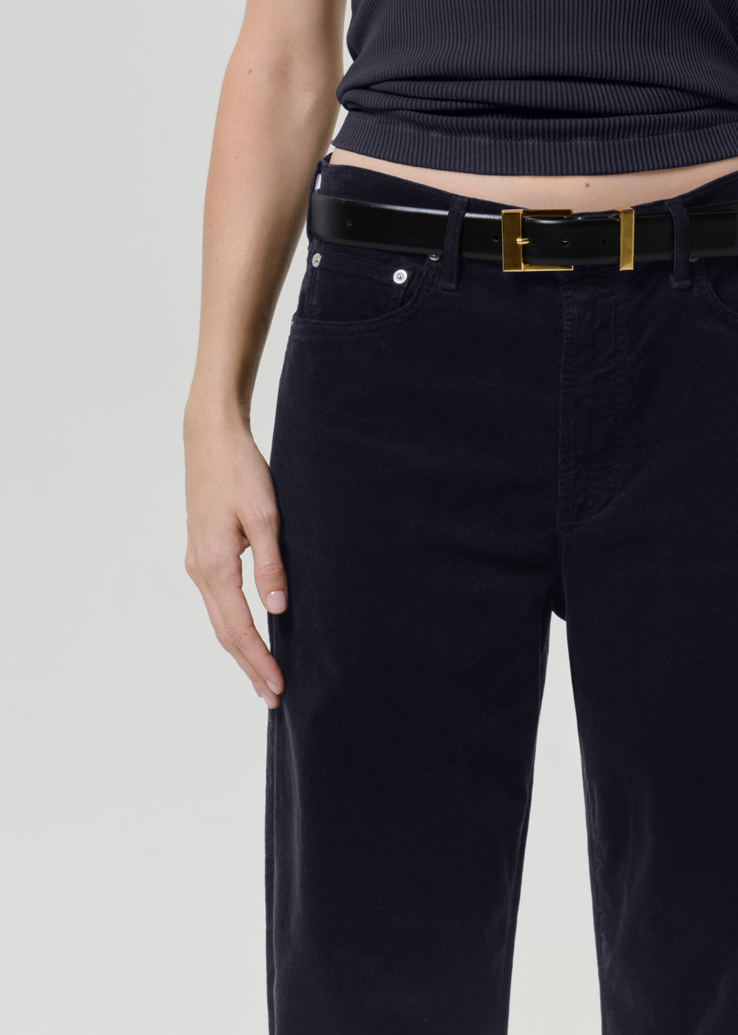 Close-up of a person wearing dark navy corduroy pants with a black belt featuring a gold rectangular buckle, paired with a dark ribbed top against a light gray studio background.