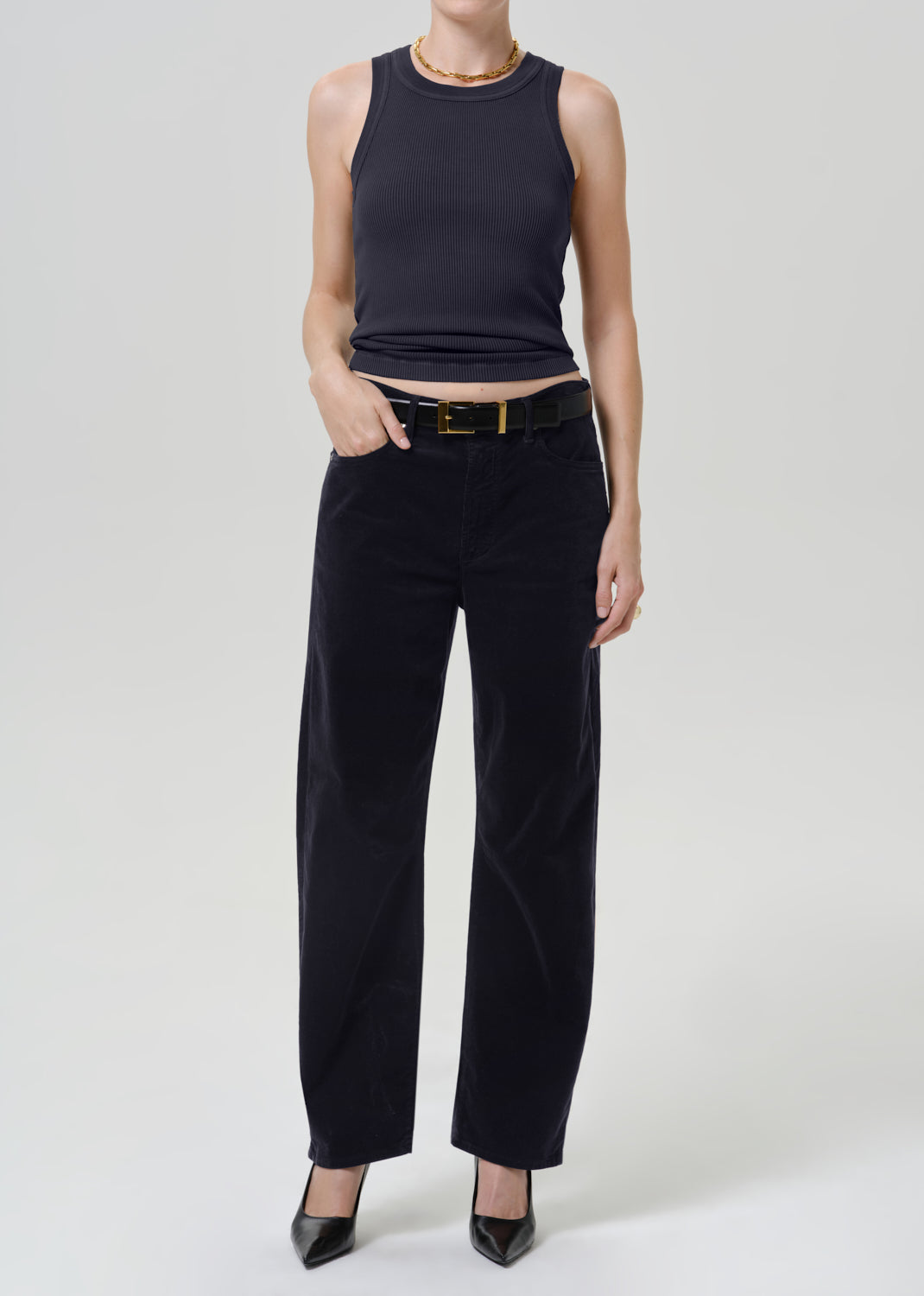 A model wearing a sleeveless black ribbed top and black wide-leg trousers with a black belt, standing against a light gray studio background.