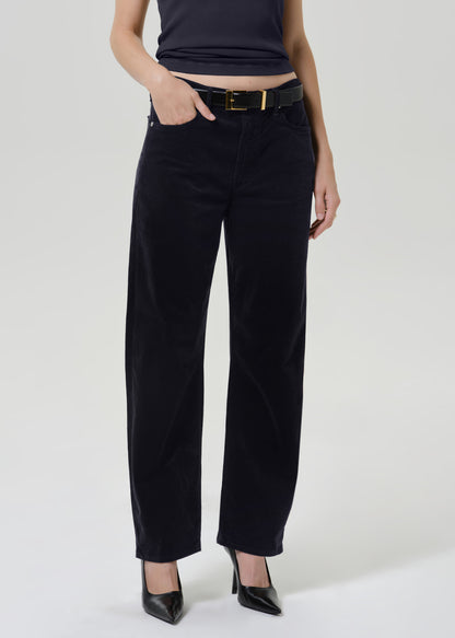 A model wearing black wide-leg corduroy trousers with a black belt and gold buckle, styled with a dark top and black pointed-toe heels against a light gray studio background.