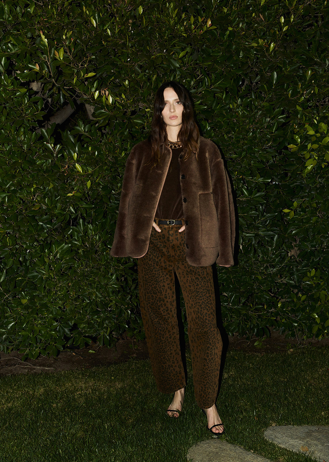 Person wearing a brown fur coat and matching pants standing against a green hedge.