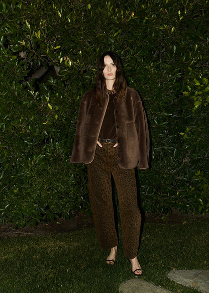 Person wearing a brown fur coat and matching pants standing against a green hedge.