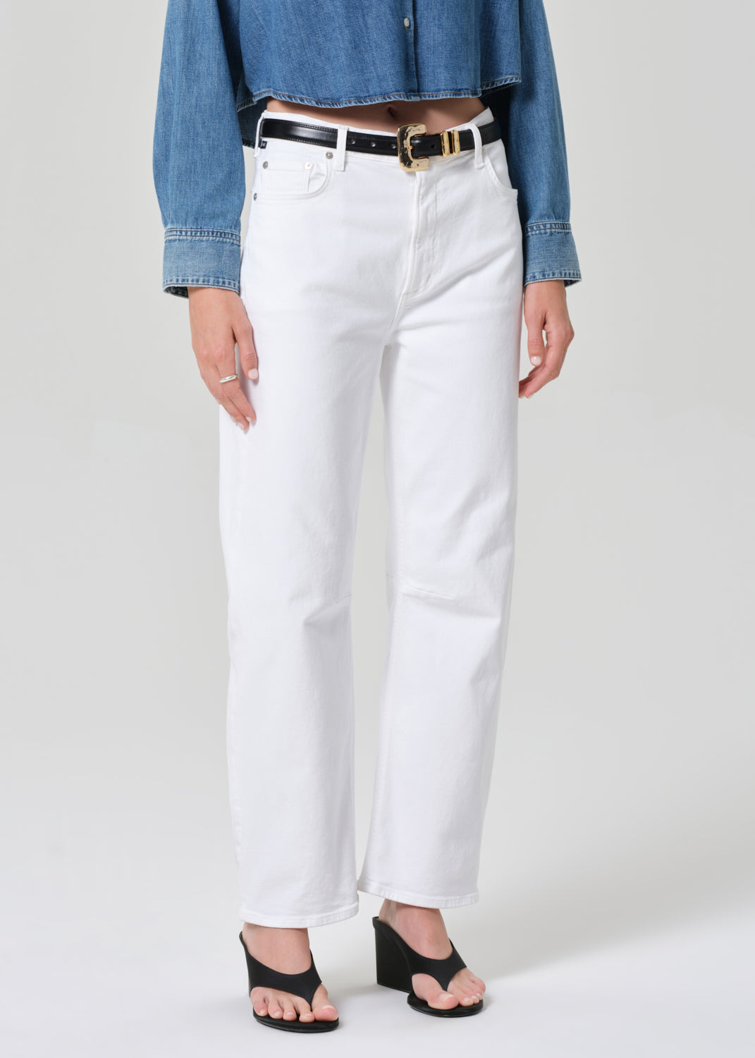 Person wearing white jeans with a denim shirt and black sandals on a plain background