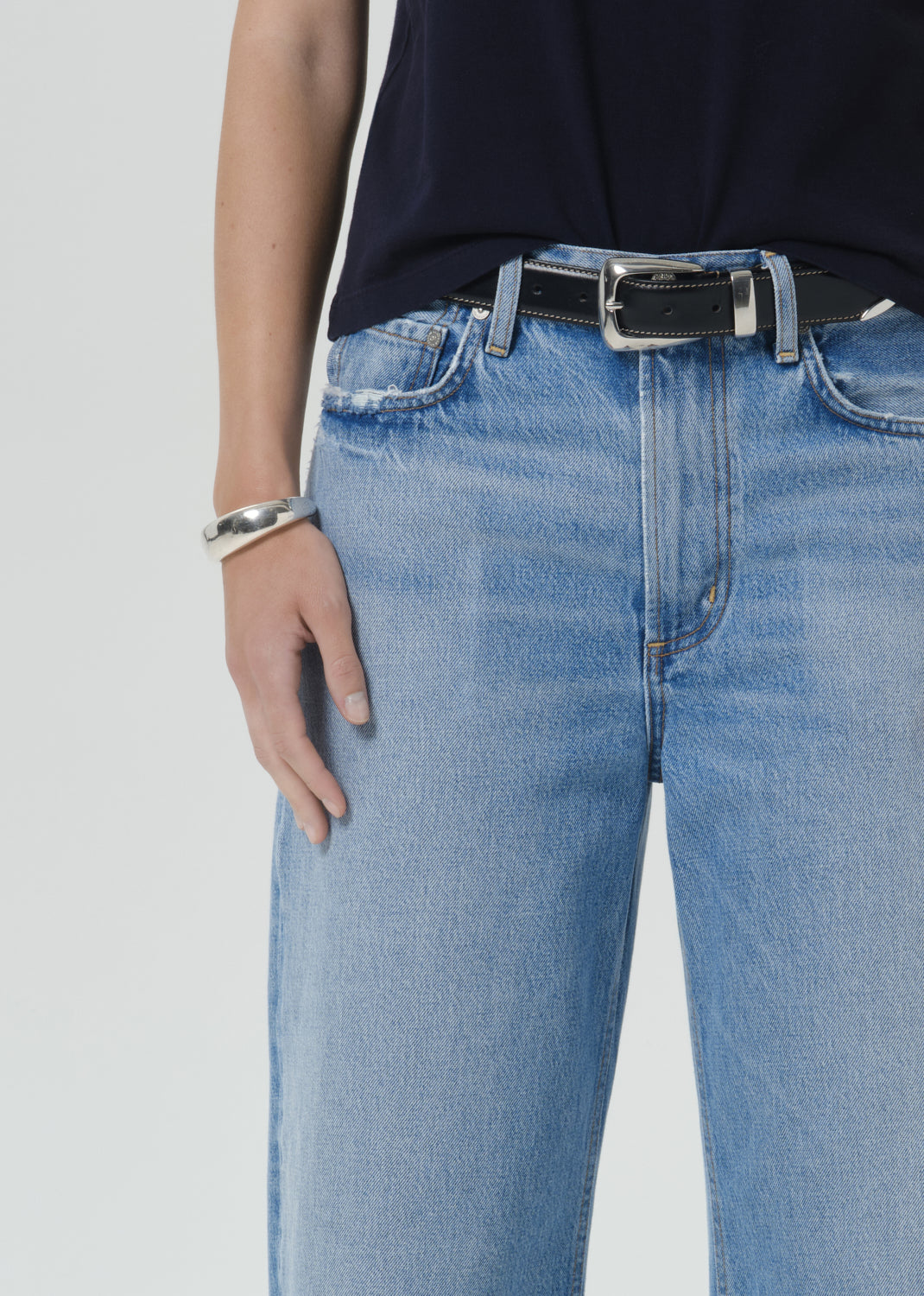 Close-up of a person wearing light blue denim jeans with a black leather belt featuring a silver buckle, paired with a dark top and a silver bracelet.