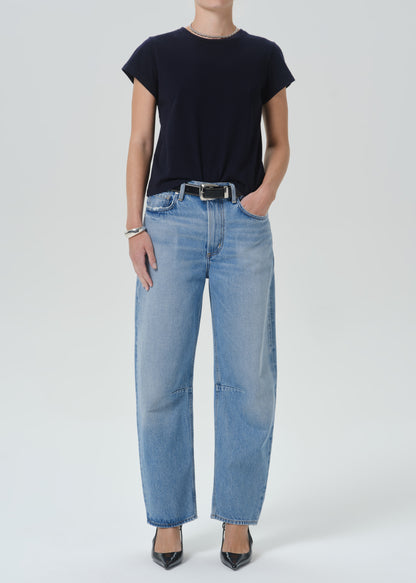 A model wearing light-wash blue wide-leg jeans with a black belt, paired with a black short-sleeve t-shirt and black pointed-toe heels, photographed against a grey background.